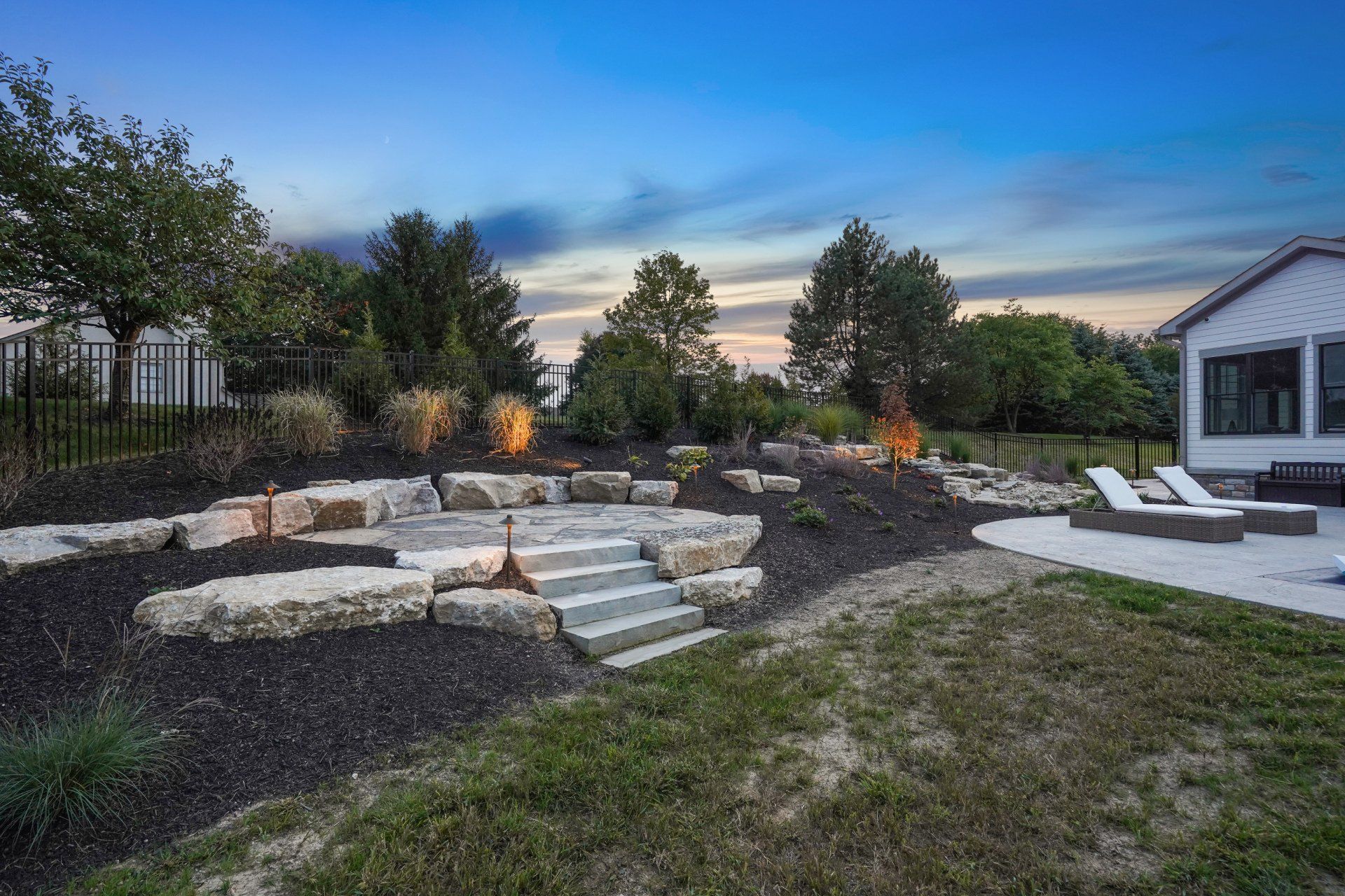 Modern backyard with stone fire pit seating, lit garden torches, and a house at dusk