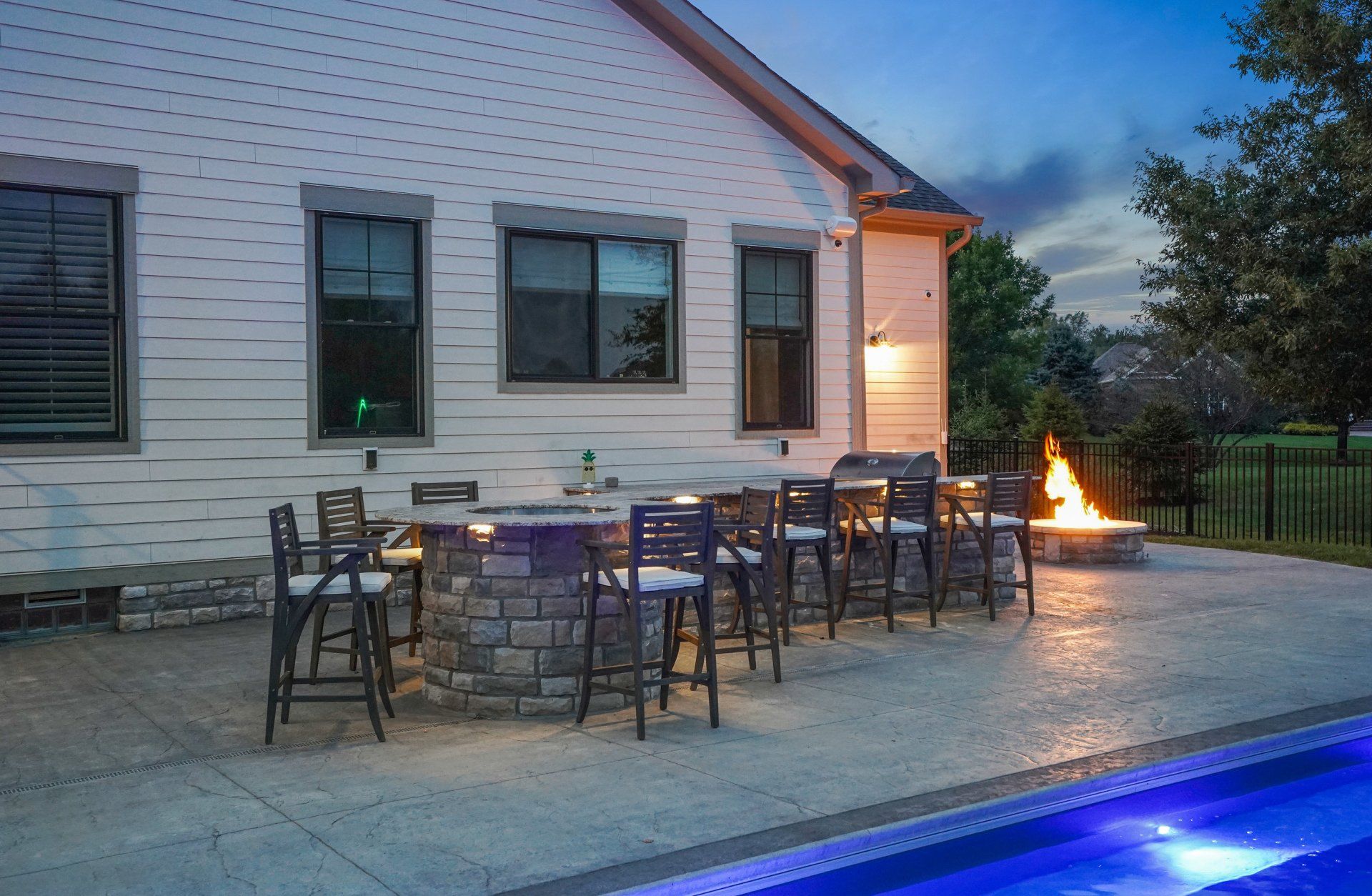 Patio dining area beside a white house at dusk, with outdoor tables, chairs, and a lit fire feature