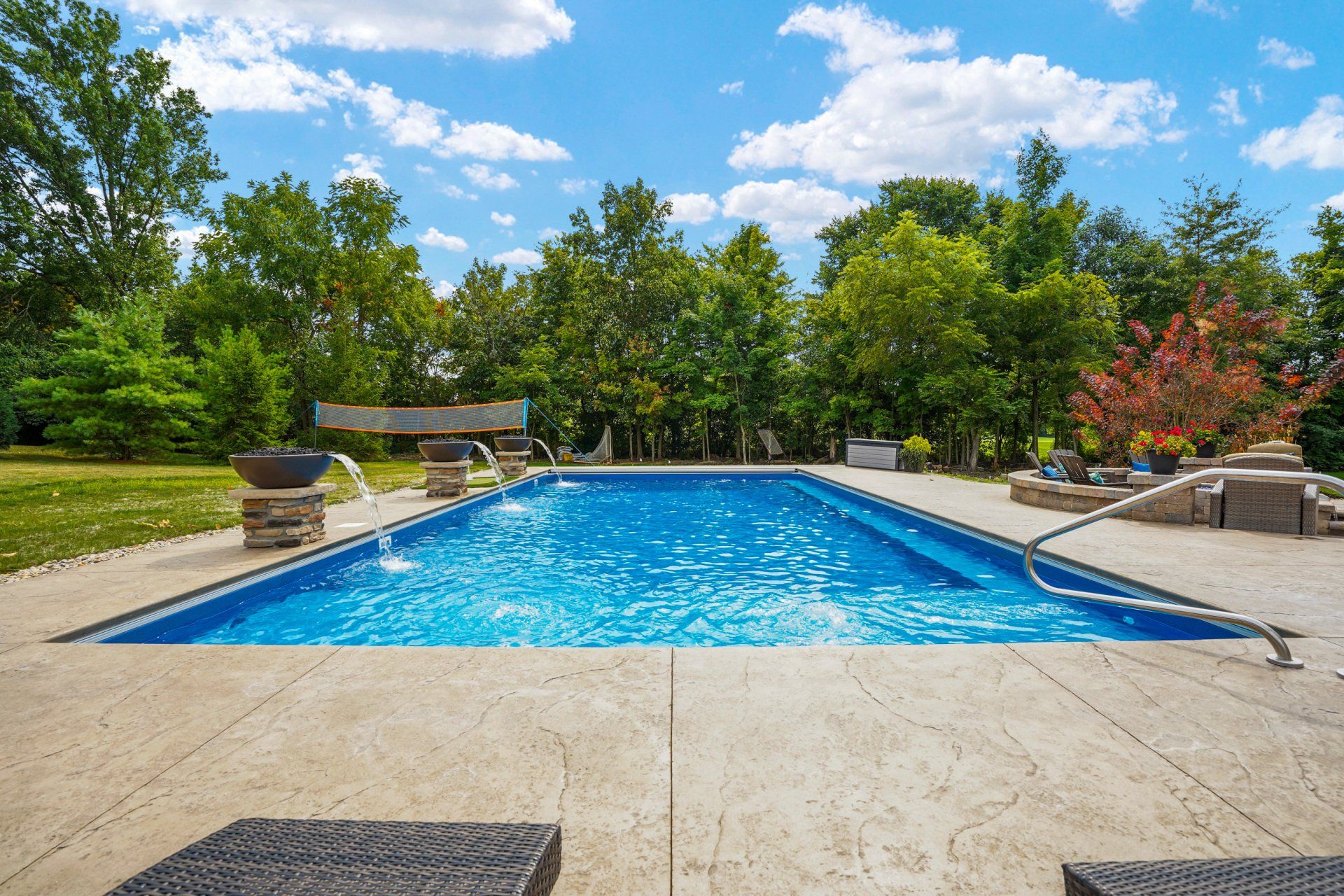Backyard swimming pool with blue water, stone patio, and lush green trees under a bright sky