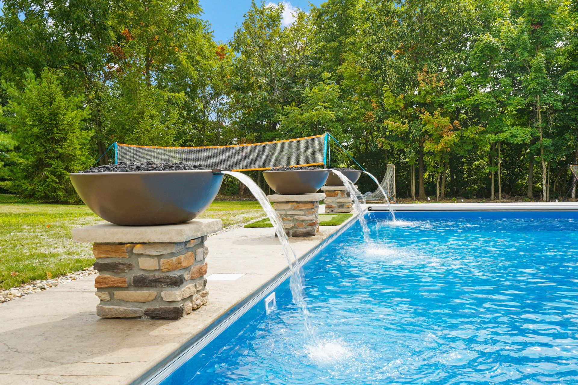 Backyard pool with a stone fire bowl and curved lounge chair beside blue water and trees
