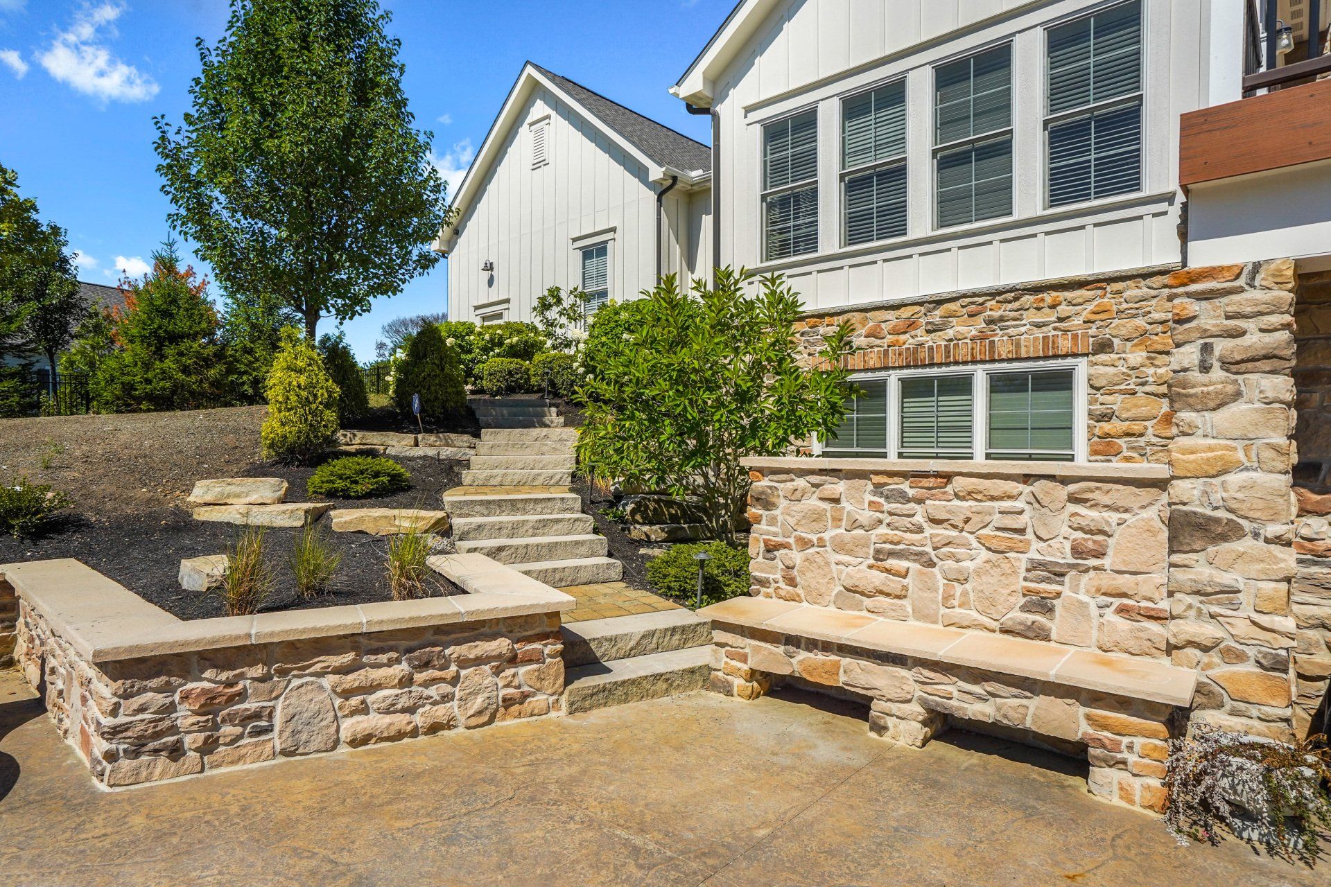 Stone patio steps and benches beside a white house with a stone wall and green shrubs