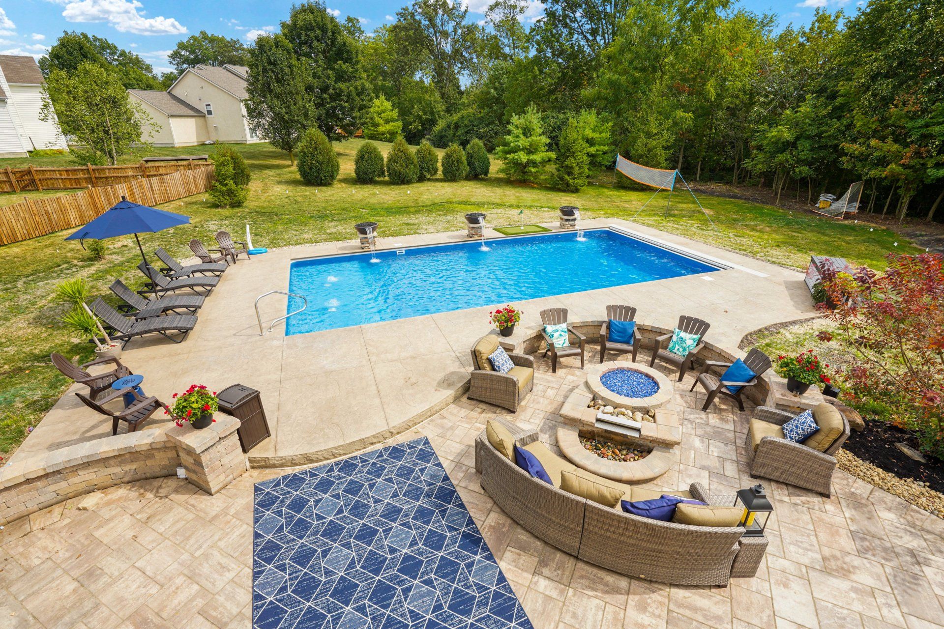 Backyard patio with blue pool, stone fire pit, lounge chairs, and green landscaping