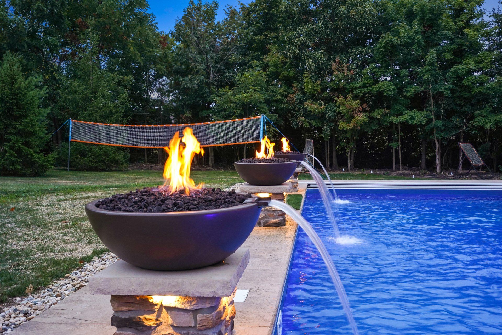 Backyard pool with fire bowls, water jets, and a volleyball net at dusk