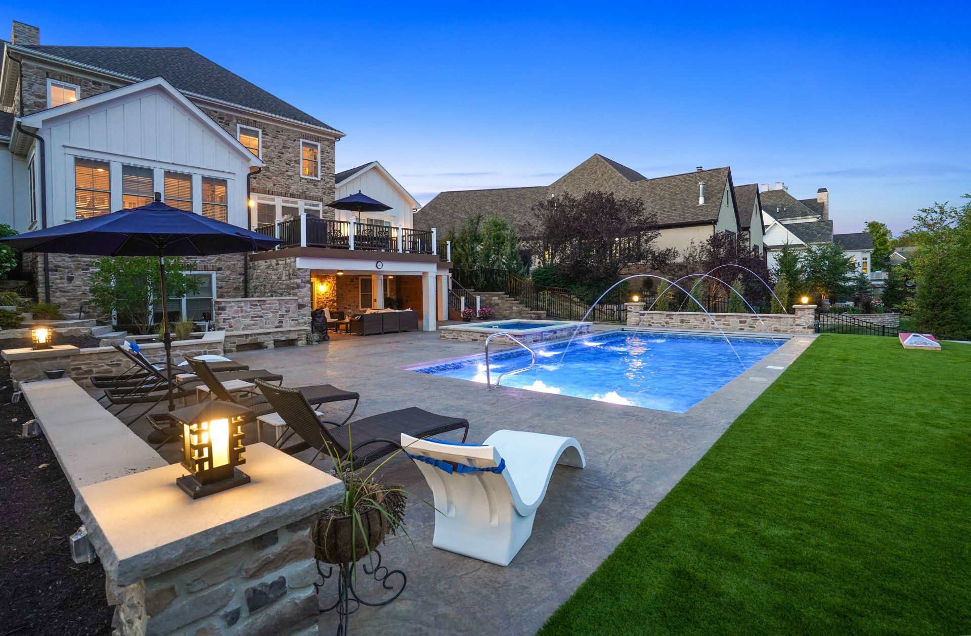 Luxury backyard with illuminated pool, stone patio seating, and a large modern house at dusk