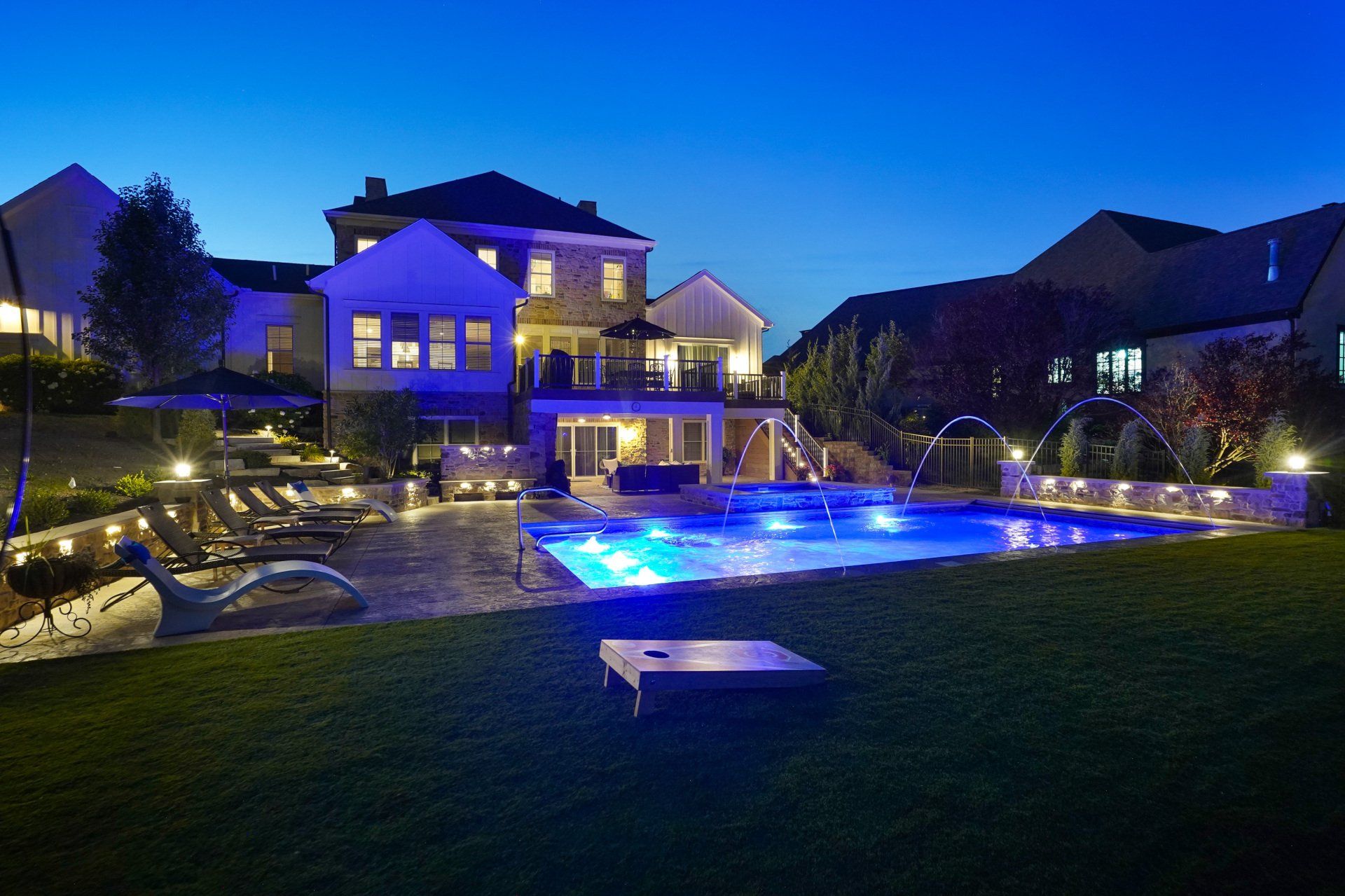 Luxury house and backyard pool lit blue at dusk, with a green lawn and neighboring homes nearby
