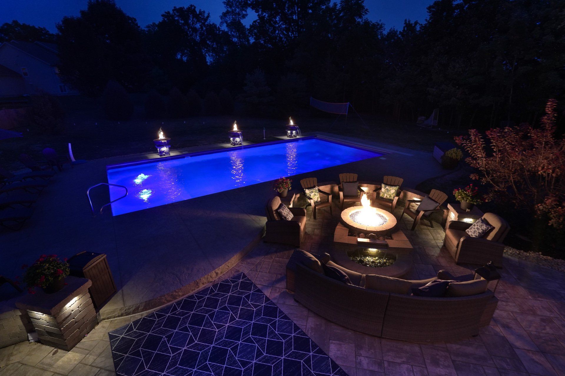 Nighttime patio with glowing blue pool, fire pit seating, and lounge chairs under dark trees