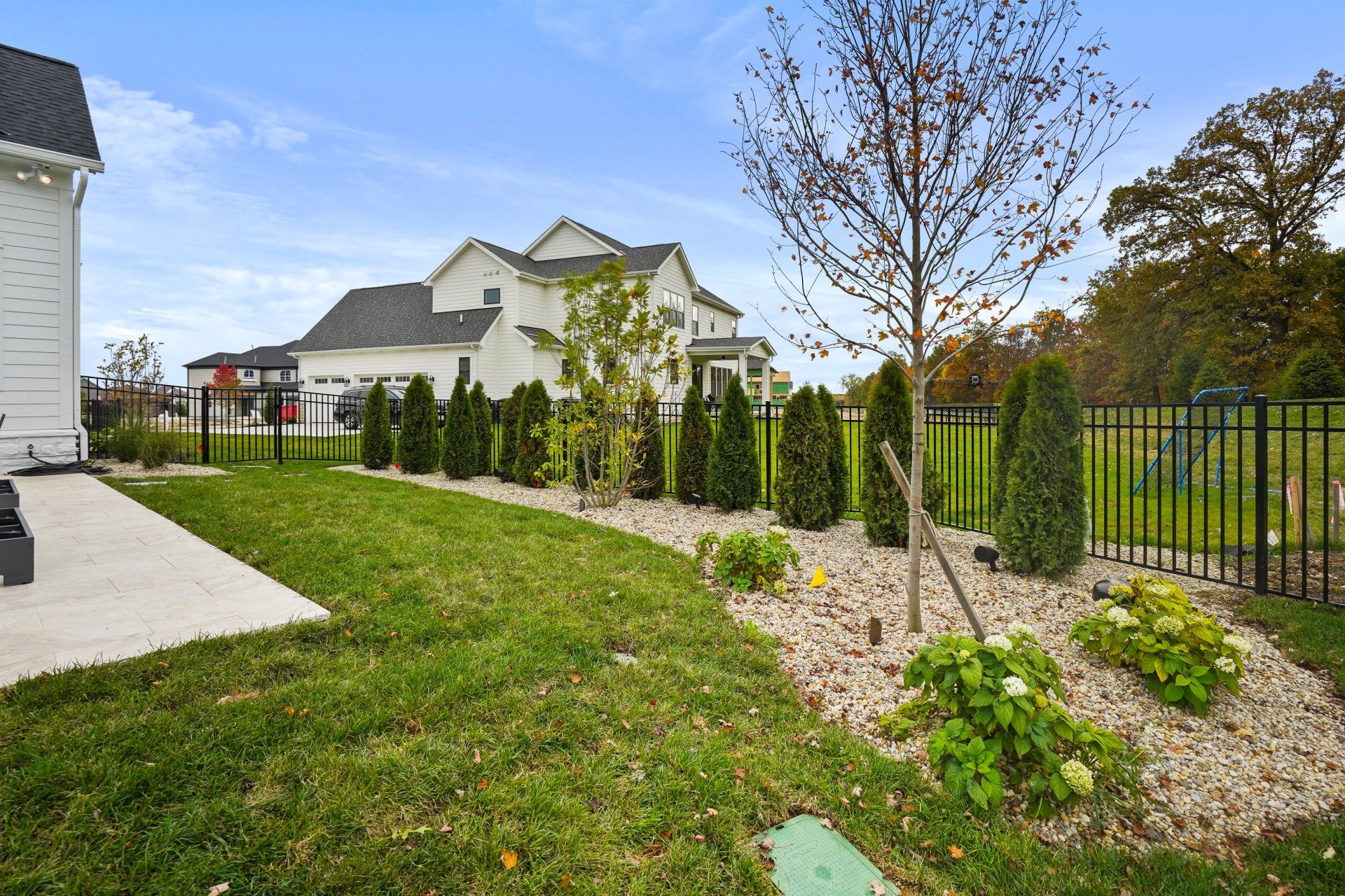 Landscaped front yard with grass, shrubs, young trees, and a black metal fence beside a house