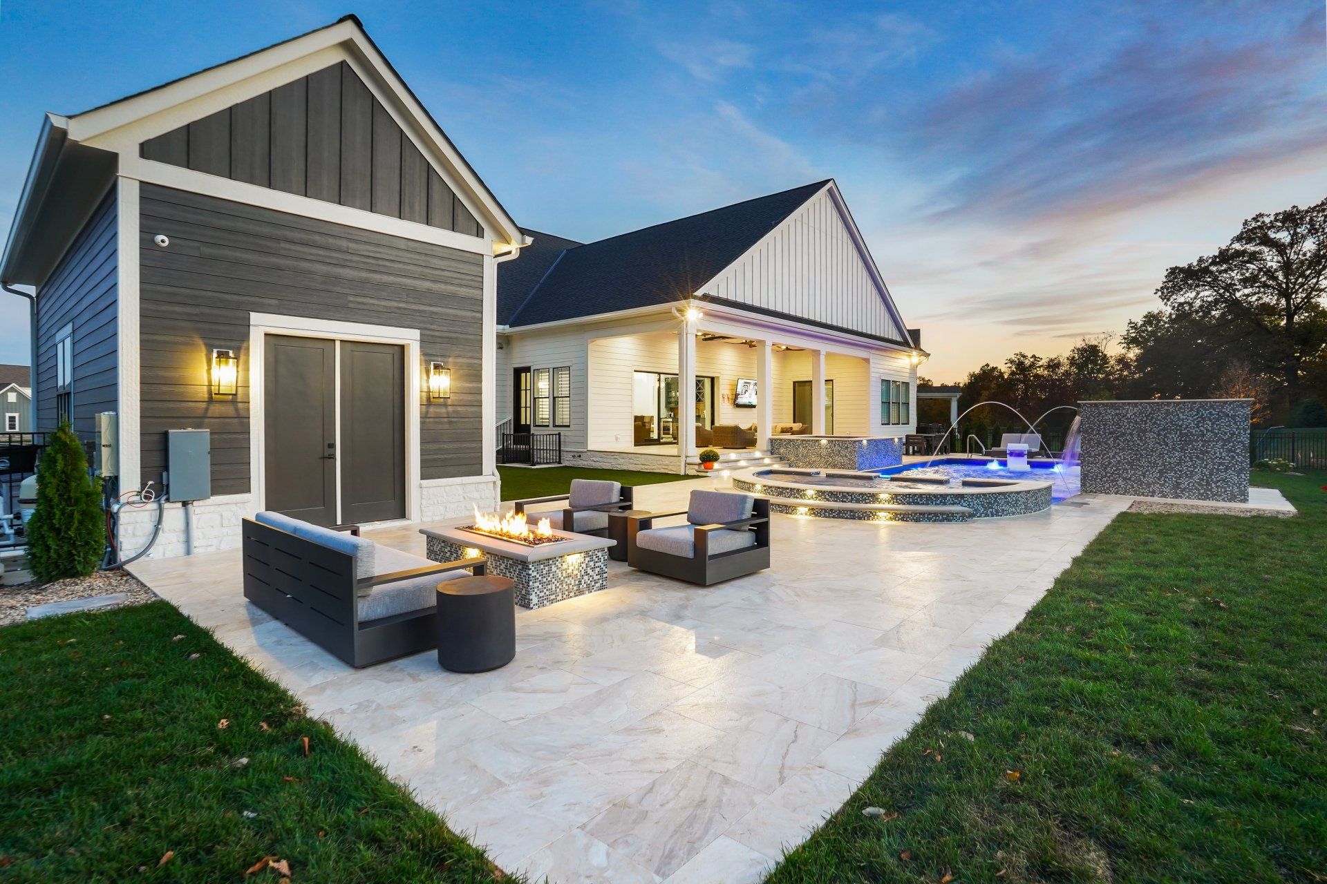 Modern backyard patio with outdoor seating, a lit fire pit, and a house exterior at dusk