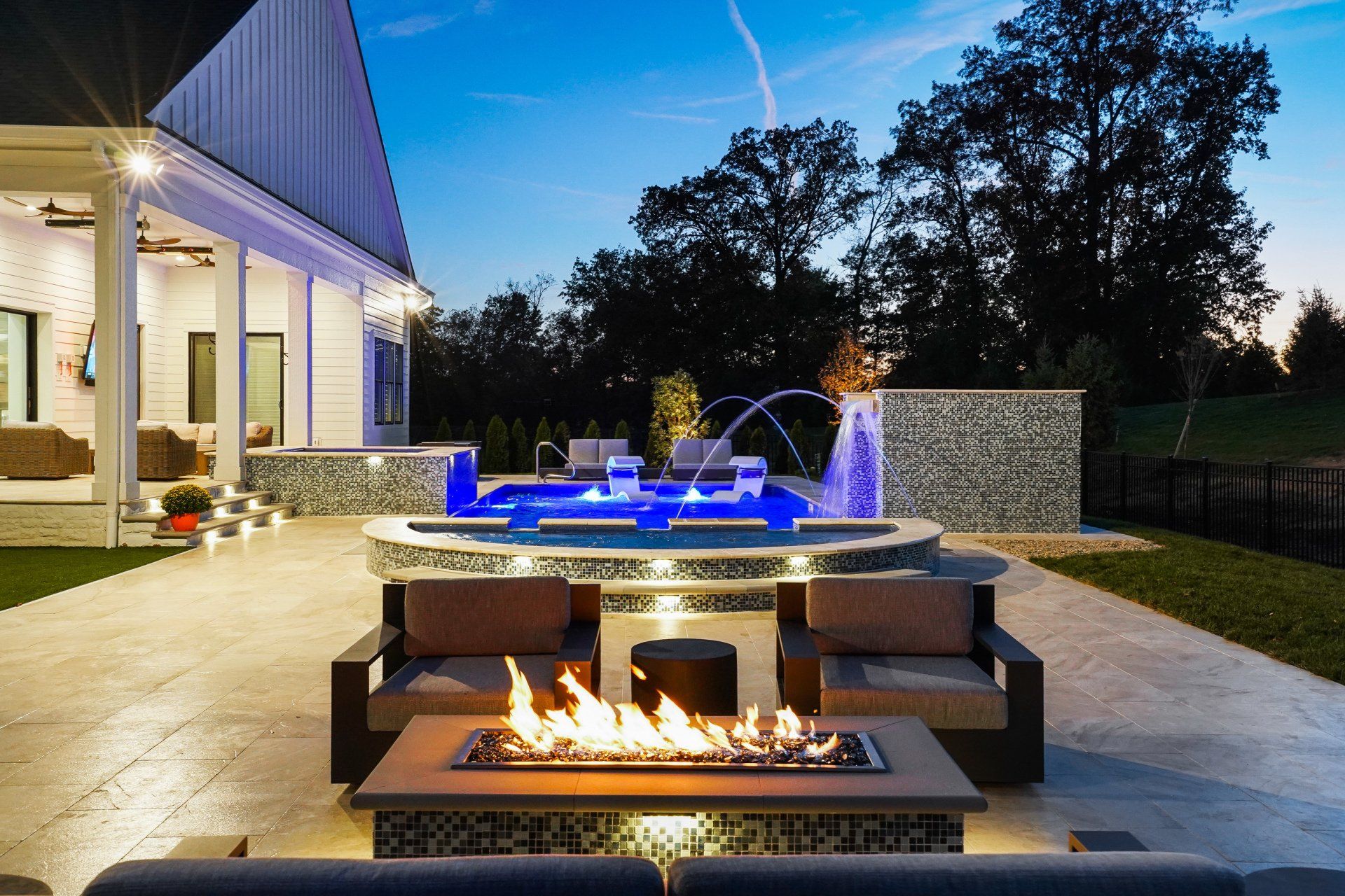Modern backyard patio at dusk with lit fire pit, blue pool, and house exterior