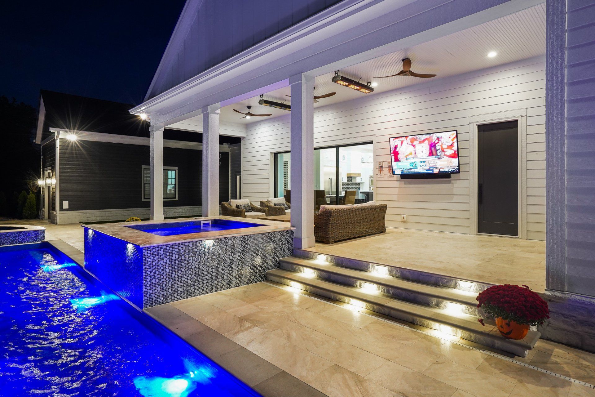 Modern patio with blue-lit pool, outdoor TV, and covered seating area at night