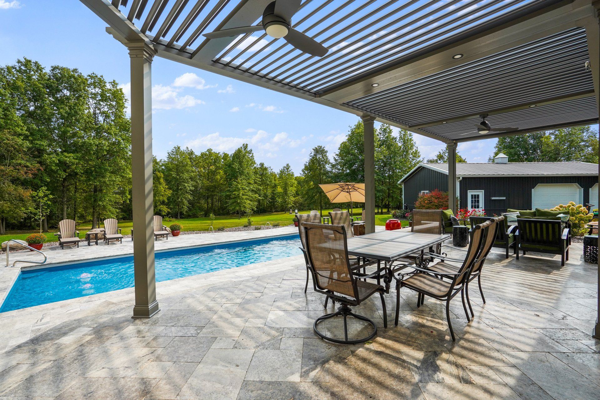 Backyard patio with dining table, pergola, and swimming pool beside lush trees and a pool house.