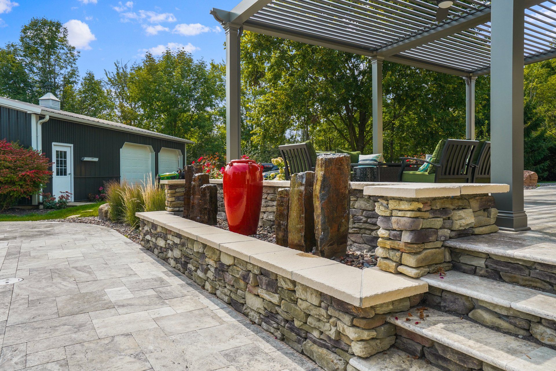 Stone patio with steps, pergola, and red urn planter beside a house and driveway