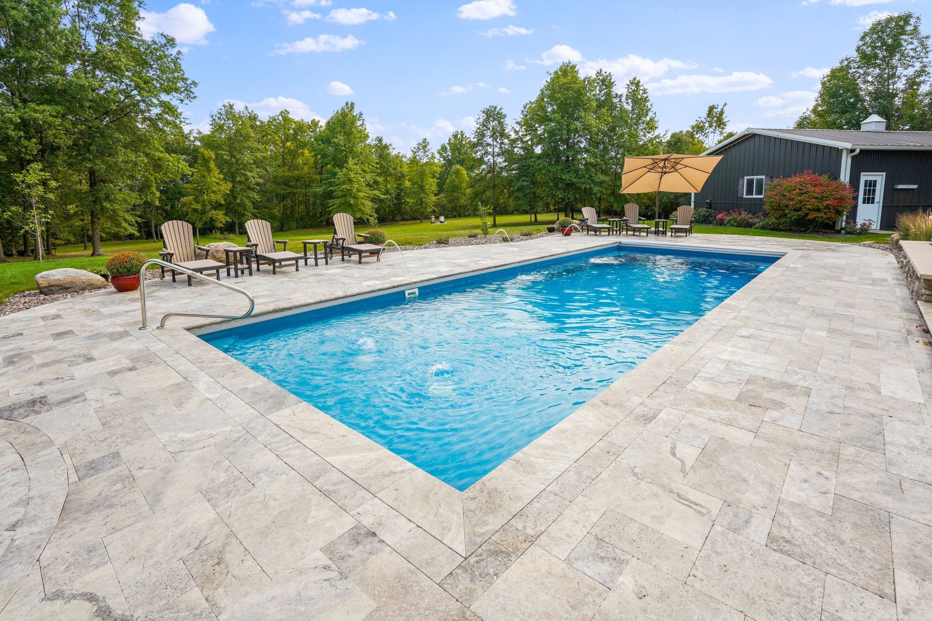 Backyard in-ground swimming pool with stone patio, lounge chairs, and trees around it