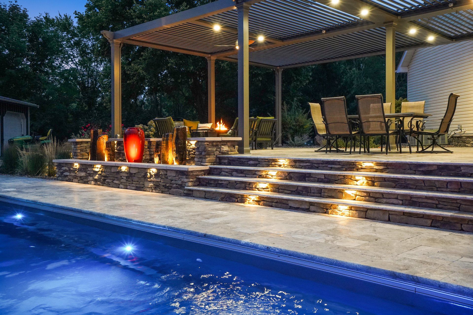 Backyard pool at dusk with lit steps, pergola seating area, and stone patio by the water