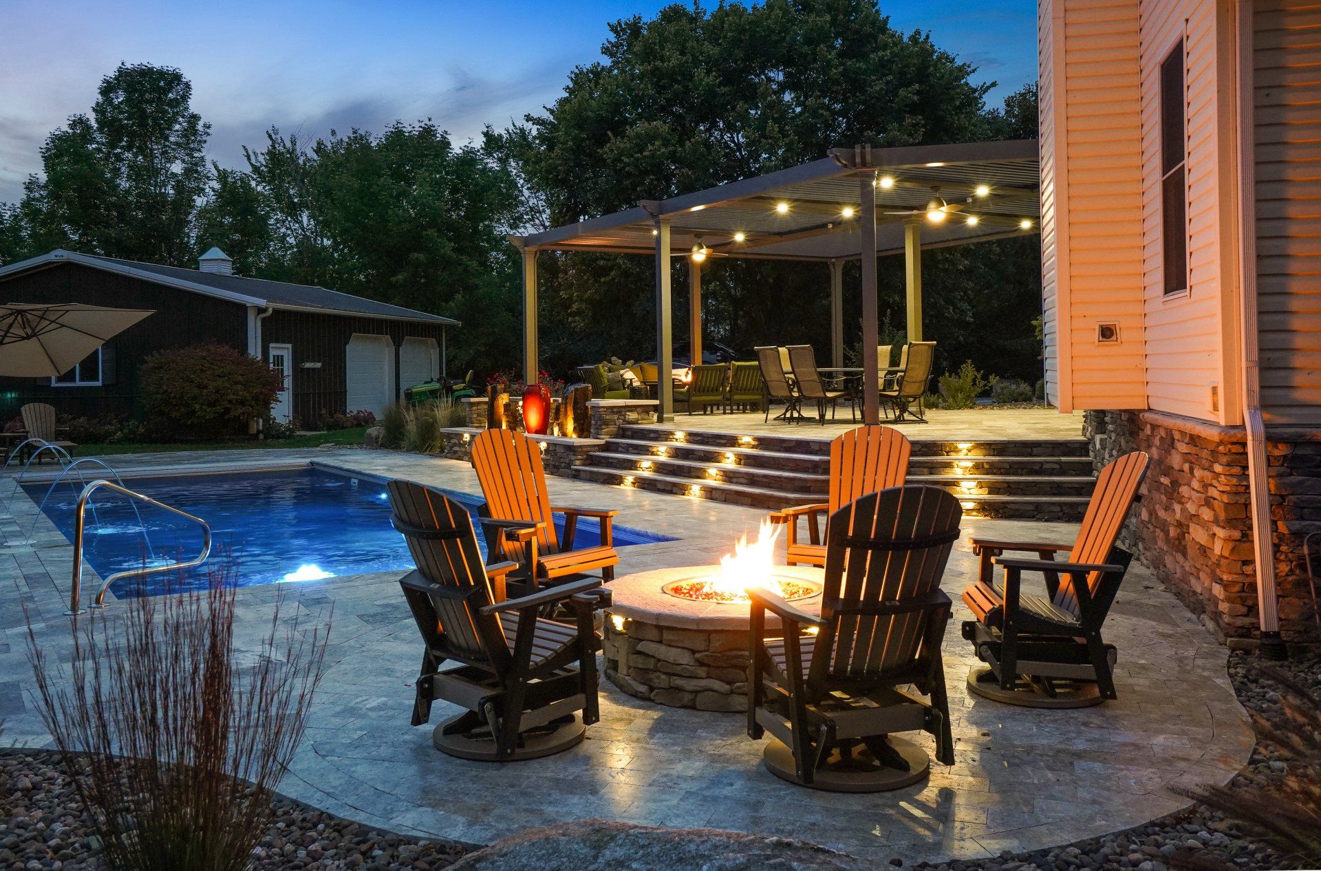 Backyard patio with fire pit, Adirondack chairs, and lit swimming pool at dusk