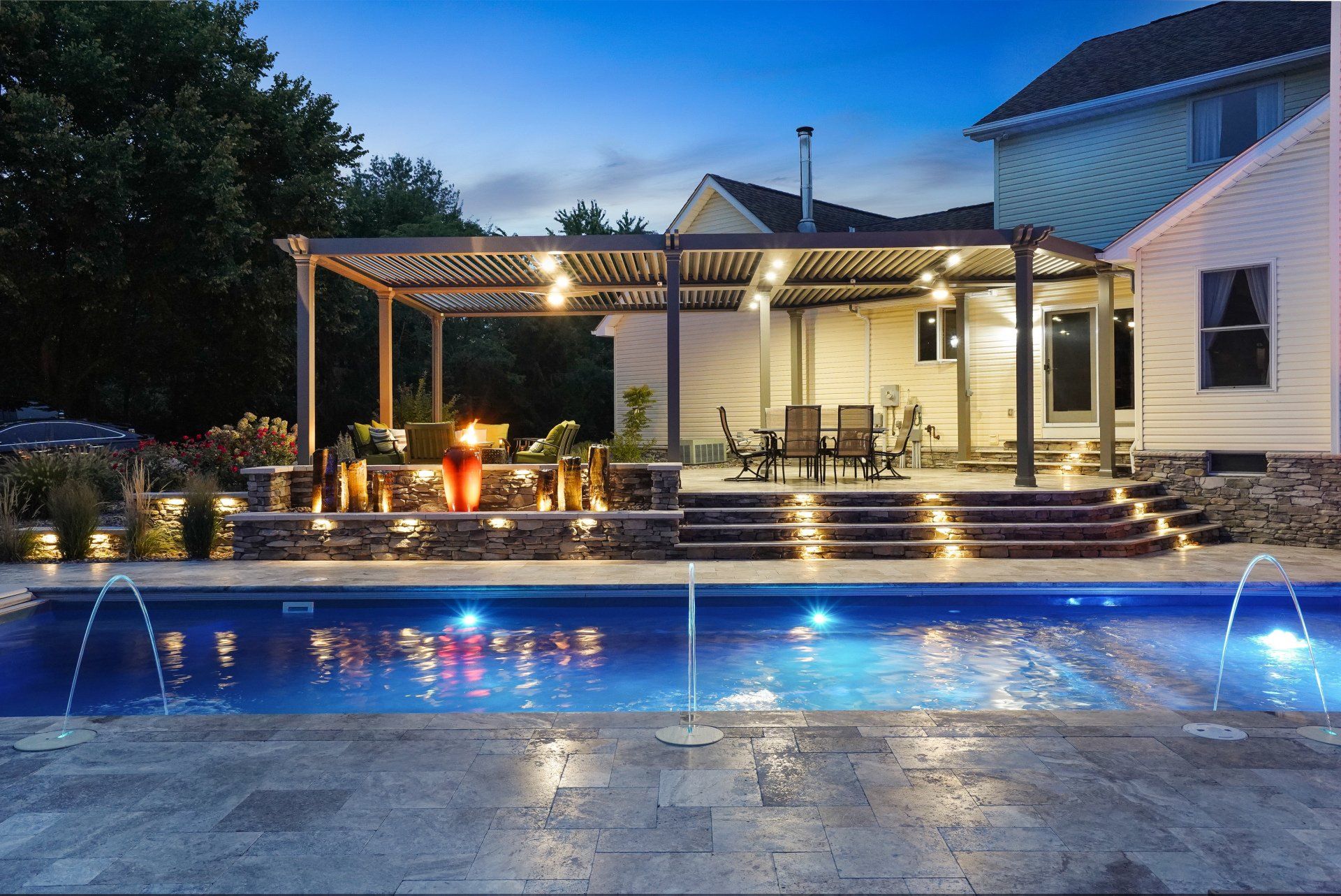 Backyard patio and lit swimming pool beside a white house at dusk