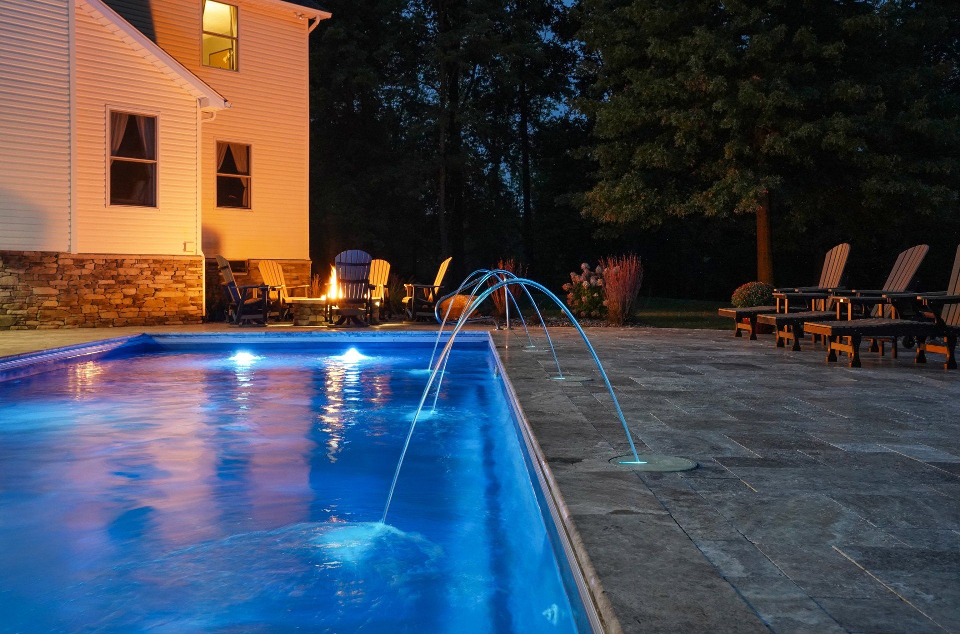 Lit backyard pool at night beside a house, with blue water jets and lounge chairs on the patio
