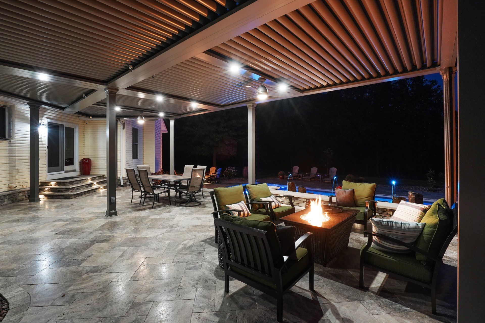 Covered patio with lounge chairs and a fire pit at night, overlooking a dark pool area