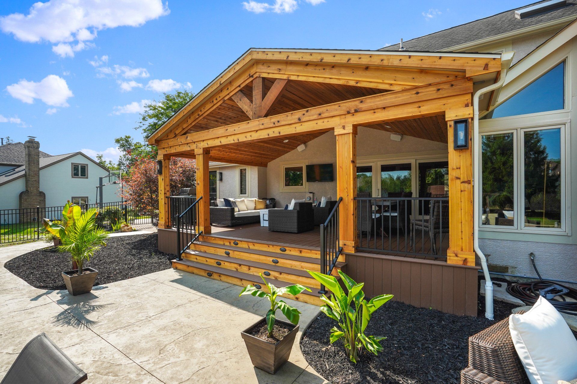 Covered wooden patio with outdoor seating, steps, potted plants, and a sunny backyard.