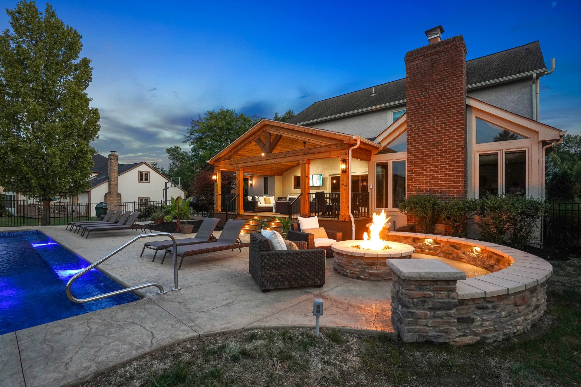 Backyard patio with lit fire pit, pool, lounge chairs, and a covered seating area at dusk