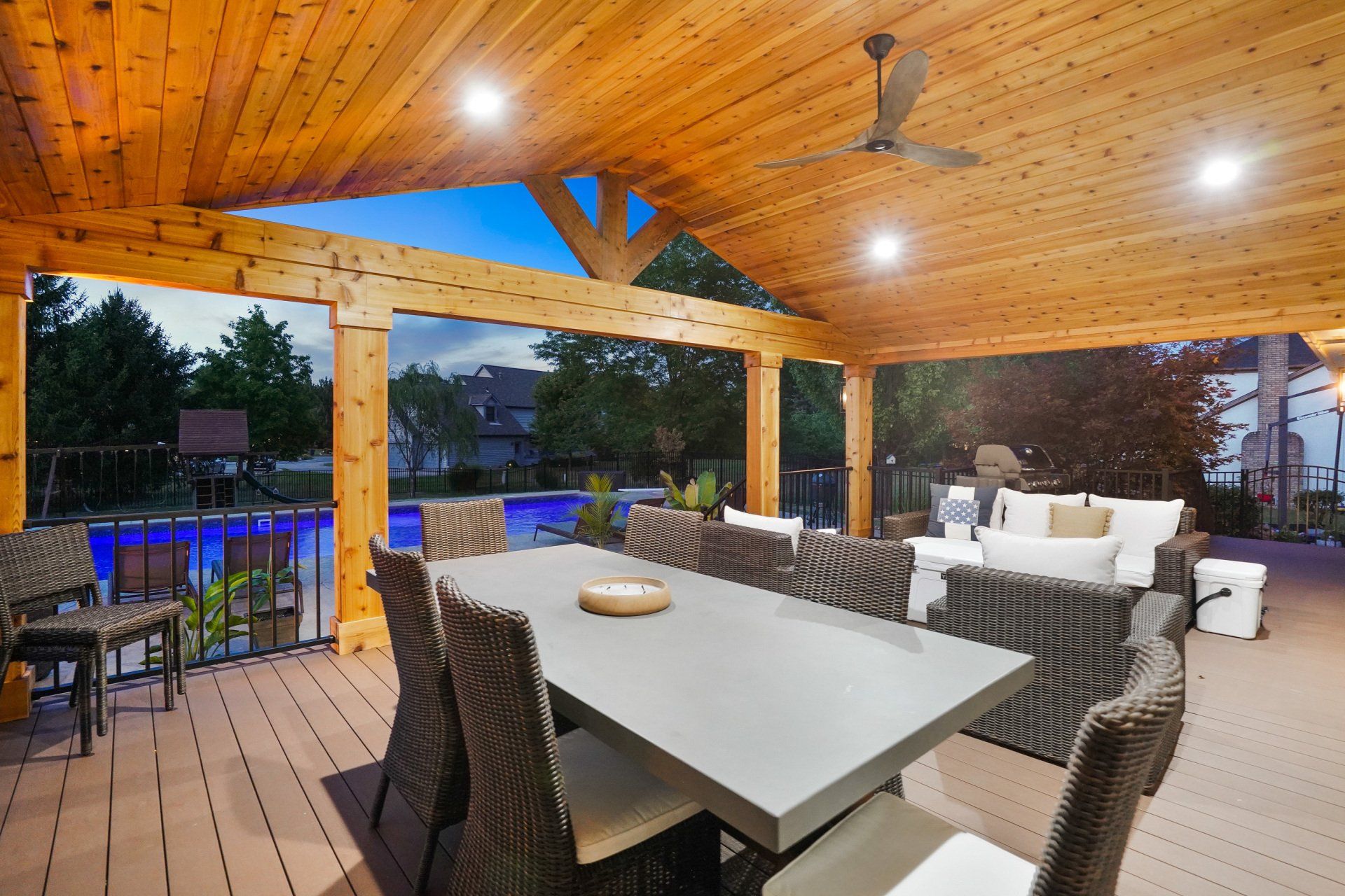 Covered outdoor patio with dining table, wicker seating, string lights, and a sunset view beyond the railing