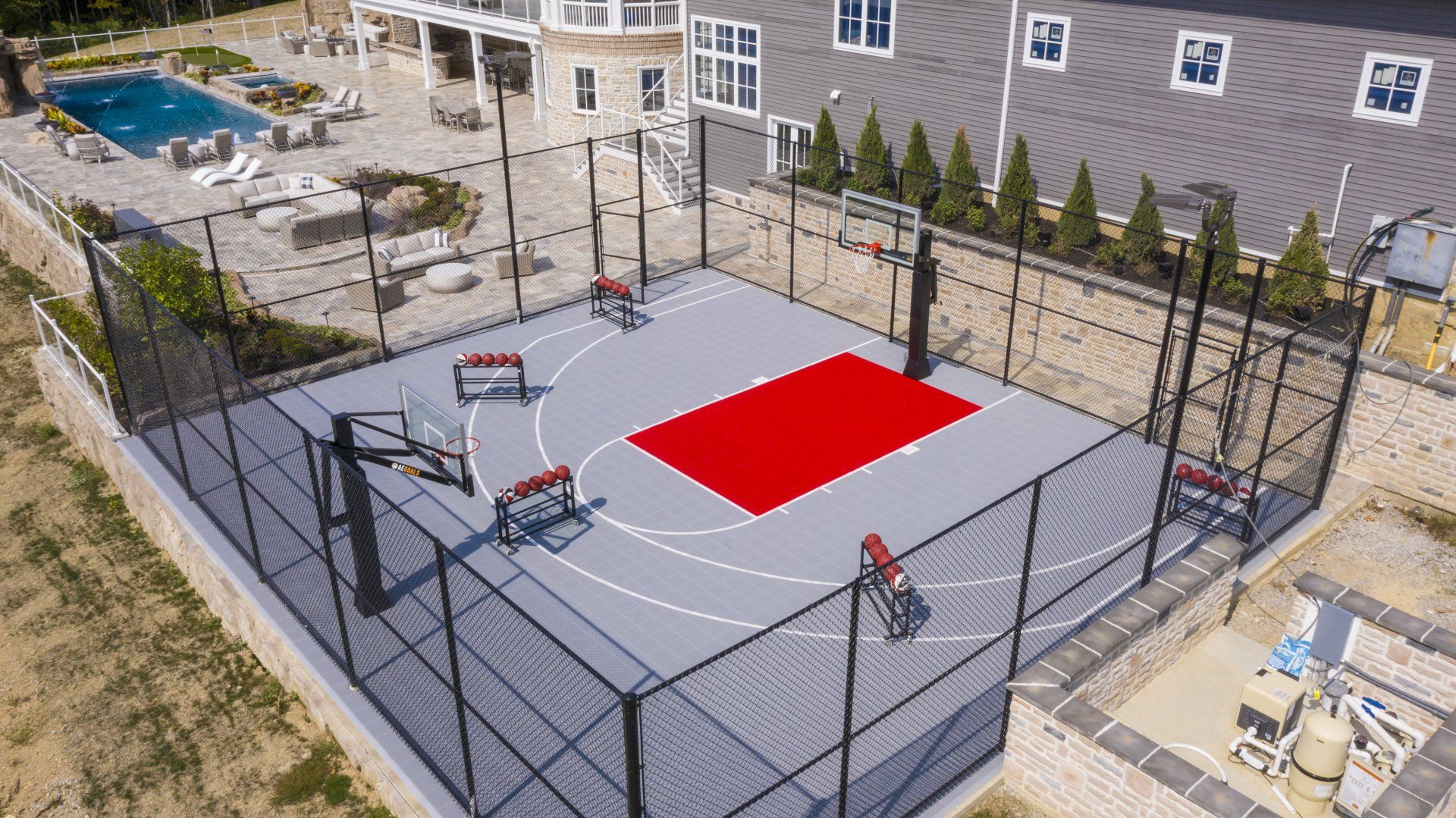 Outdoor fenced sports court with gray surface, red center rectangle, and poolside homes in the background