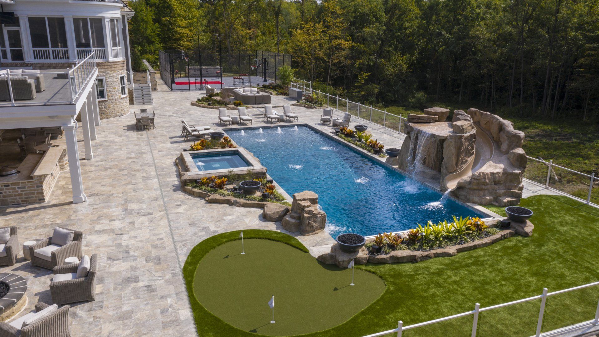 Backyard pool with blue water, stone patio, waterfall feature, and house deck surrounded by trees