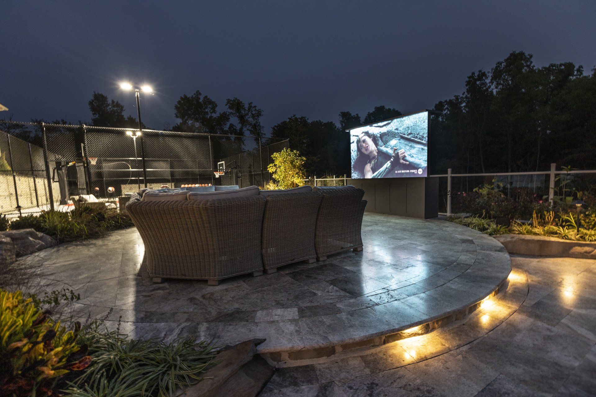 Outdoor patio at dusk with woven seating, lit steps, and a large screen showing a movie