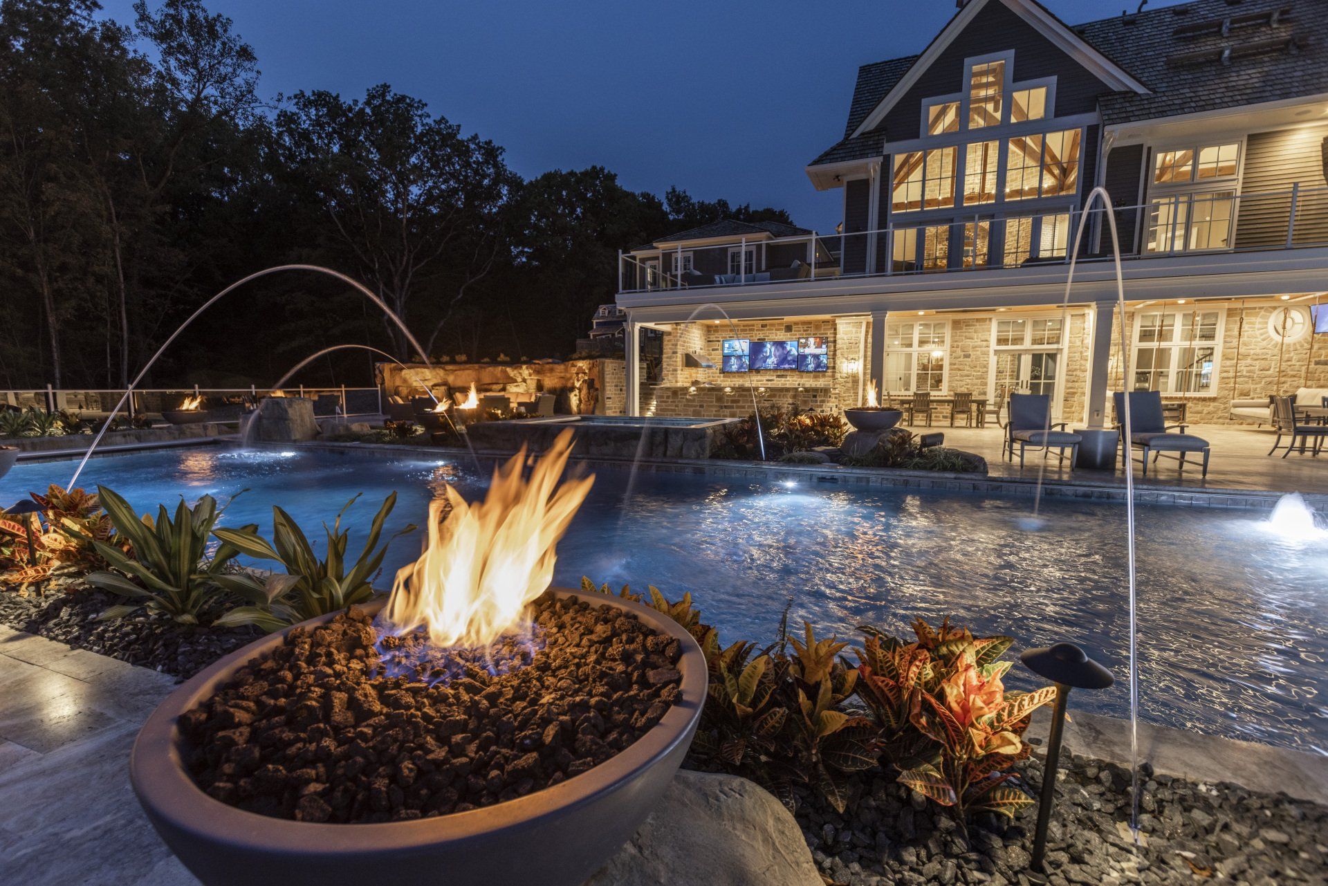 Lit fire pit by a resort pool at dusk, with a lit hotel building and fountains in the background