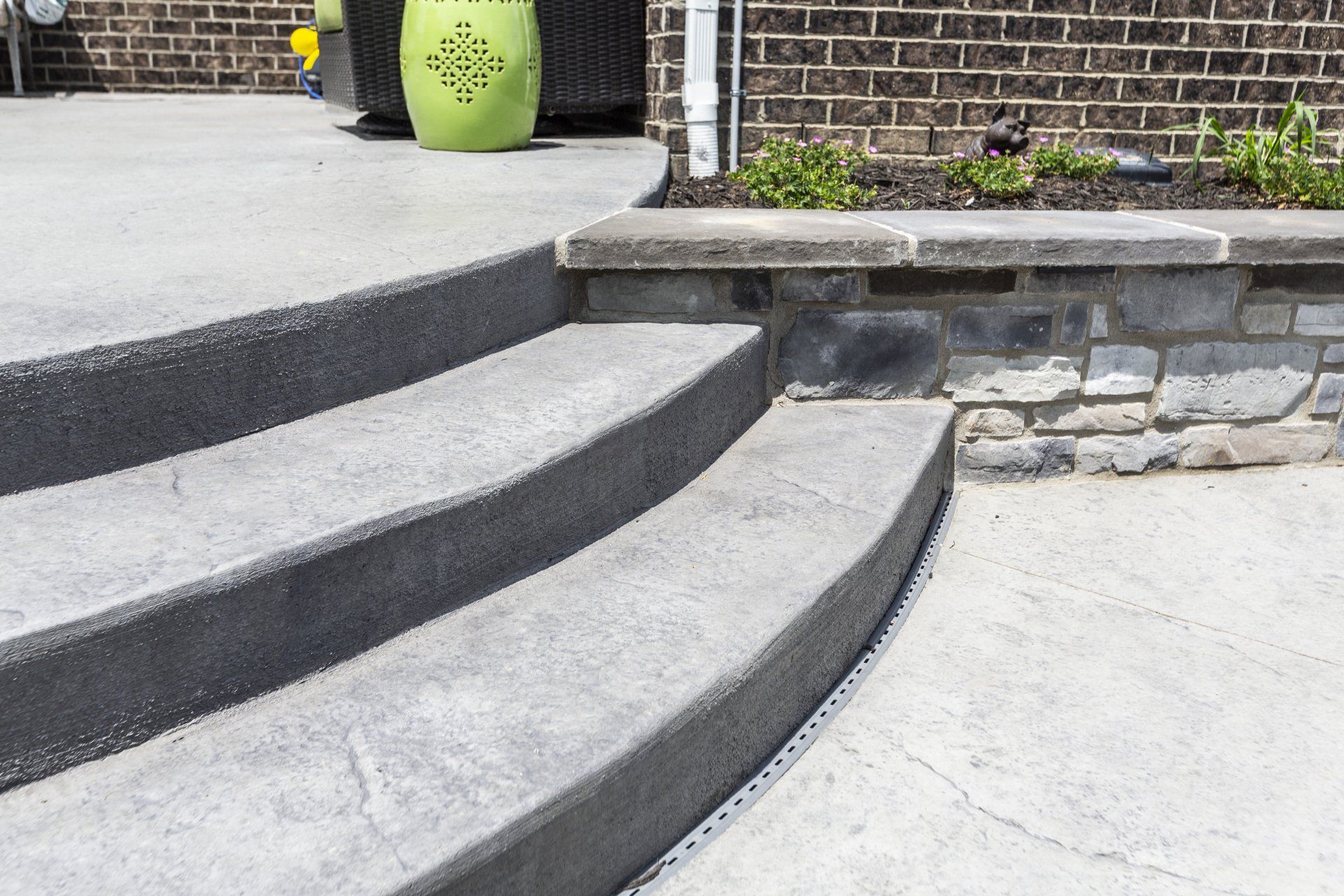 Concrete front steps with dark curved edges leading to a raised patio and stone retaining wall.