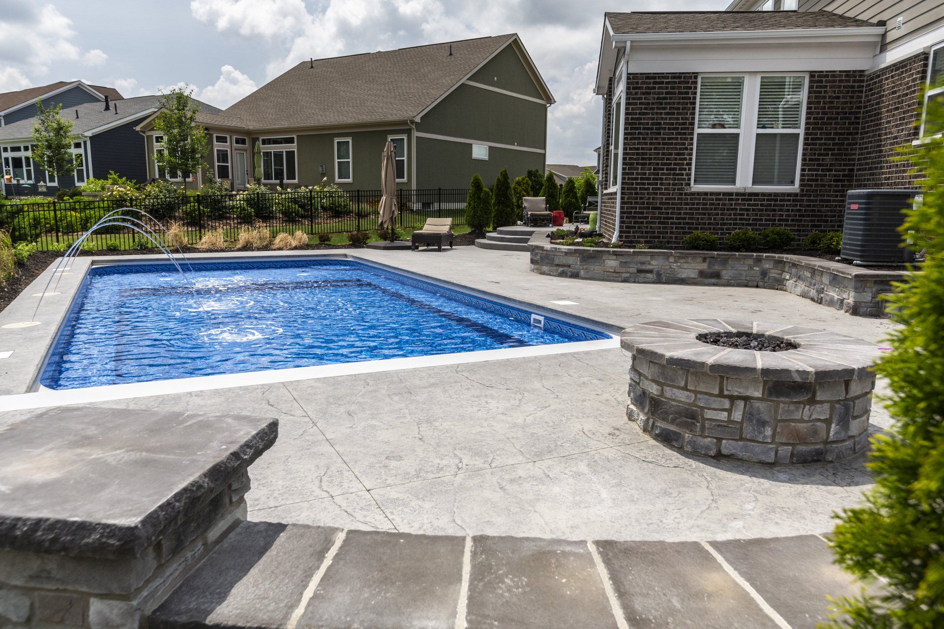 Backyard patio with a rectangular blue swimming pool and stone fire pit beside a house