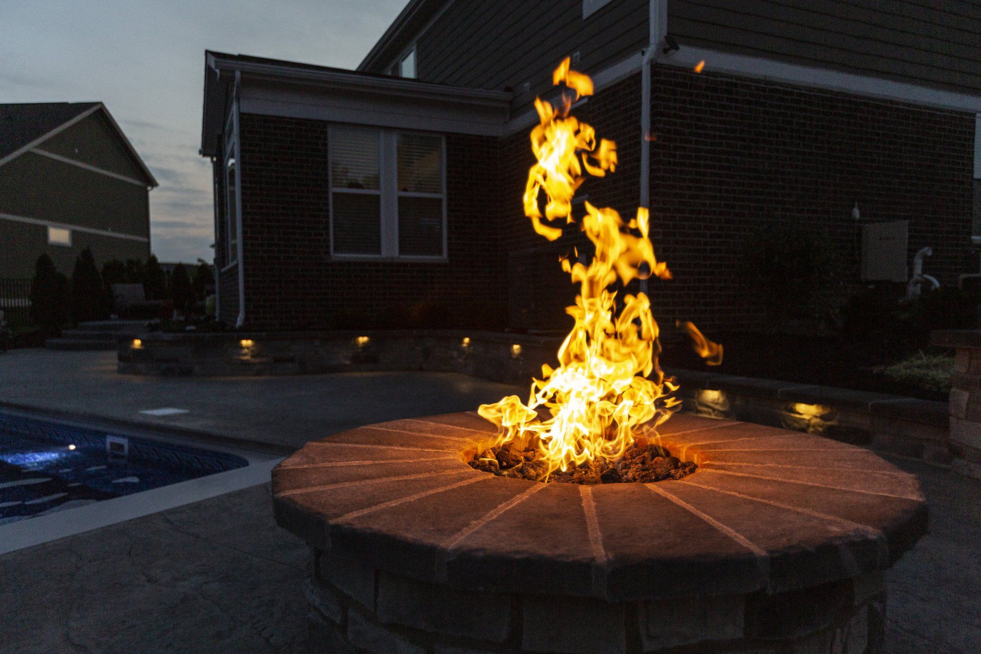 Flames rising from a circular stone fire pit in a dark suburban yard at dusk