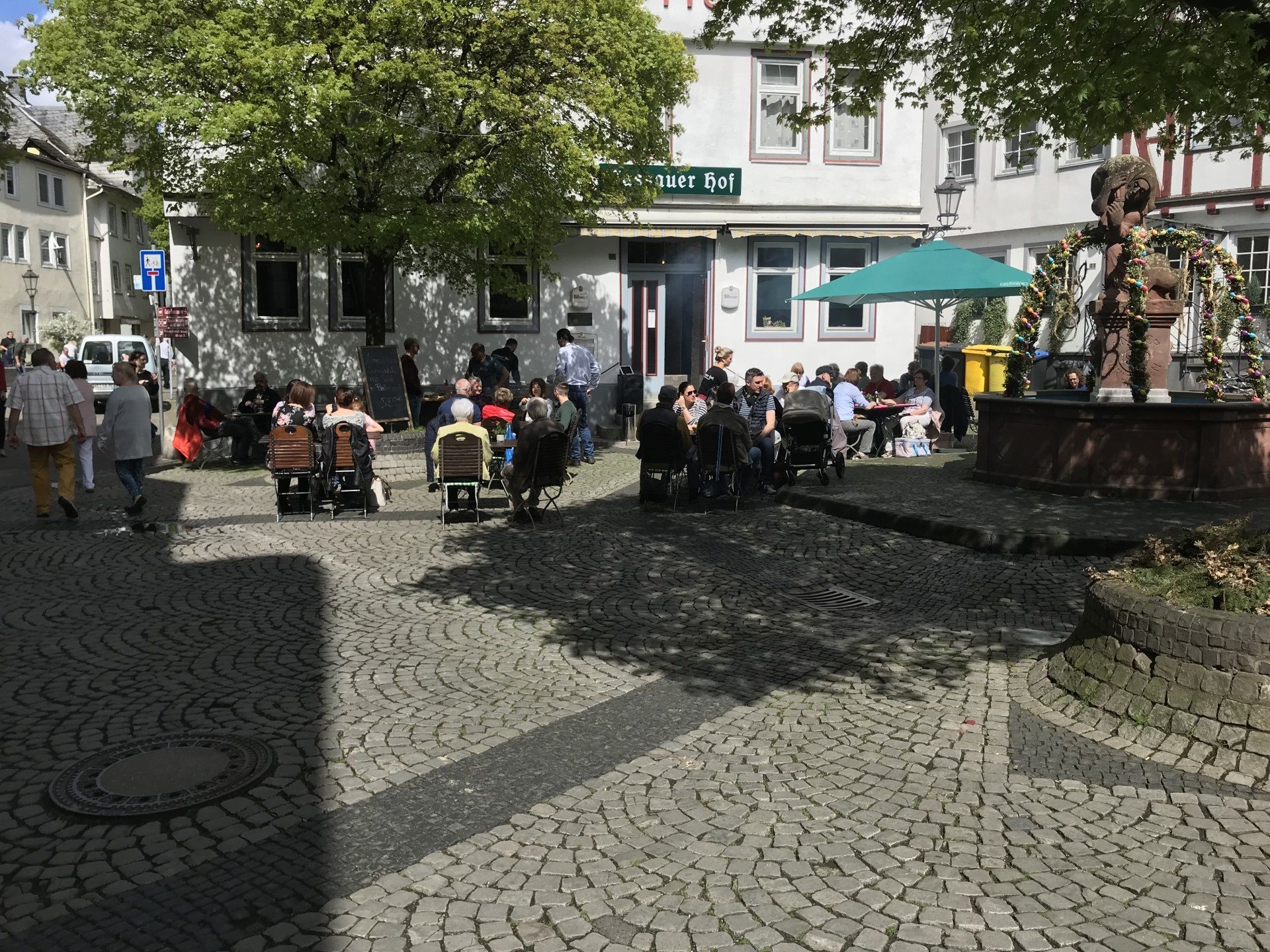 Eine Gruppe von Menschen sitzt an Tischen auf einem gepflasterten Platz vor einem Gebäude.