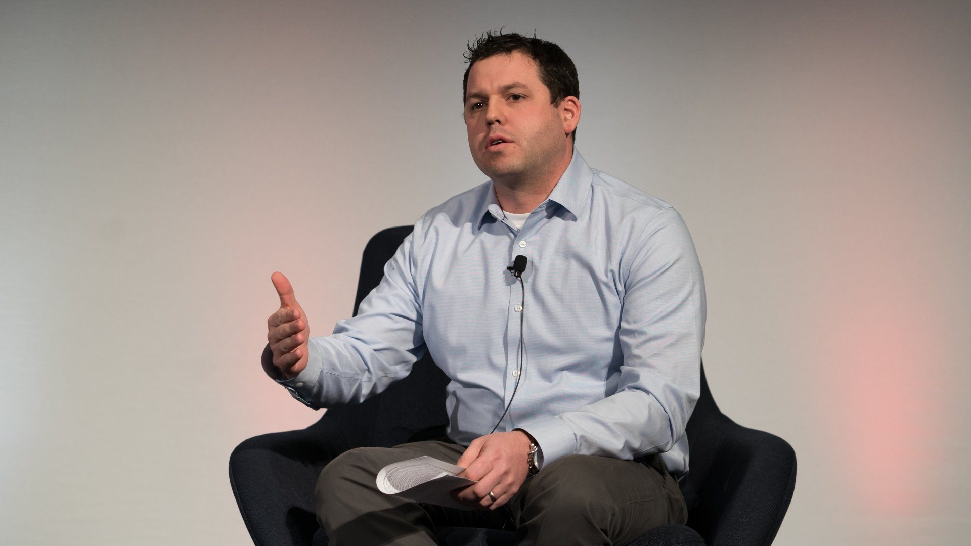 A man in a blue shirt is sitting in a chair giving a speech.