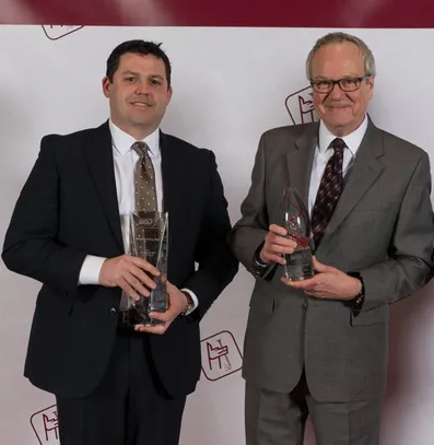 Two men in suits and ties are holding trophies