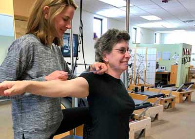 A woman is helping an older woman stretch her arms in a gym.