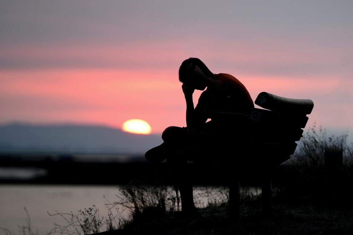 Silhouette of a person sitting on a bench, head in hand, at sunset over water.