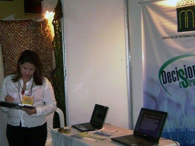 Woman at a booth with laptops and sign. She holds a clipboard, white shirt, black pants.