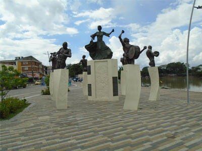 Statues of musicians and dancer on stone pedestals in a park; river and buildings in background.