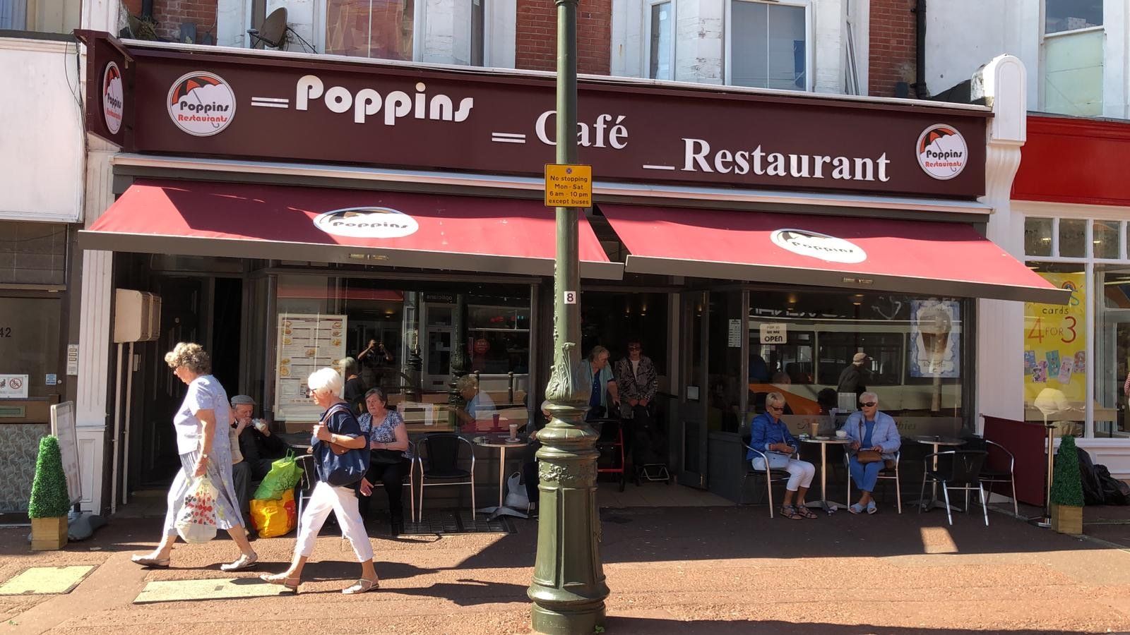 Outdoor restaurant storefront with red awnings and people seated at tables on a sunny sidewalk.