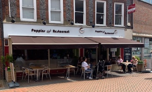 Outdoor café seating at Poppies Restaurant on a brick street, with people dining under a brown awning.