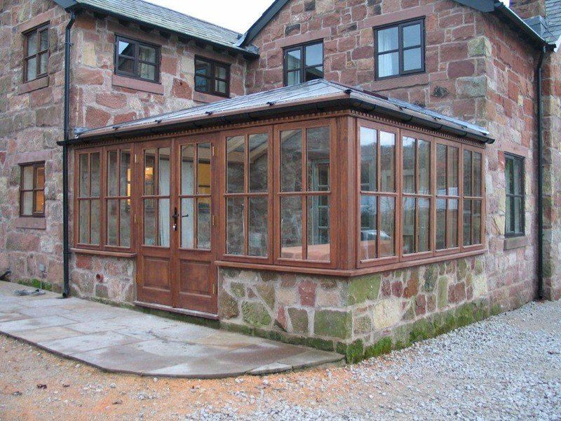 A sunroom with wooden frames and doors adjoins a red stone house. A stone patio leads up to the sunroom.