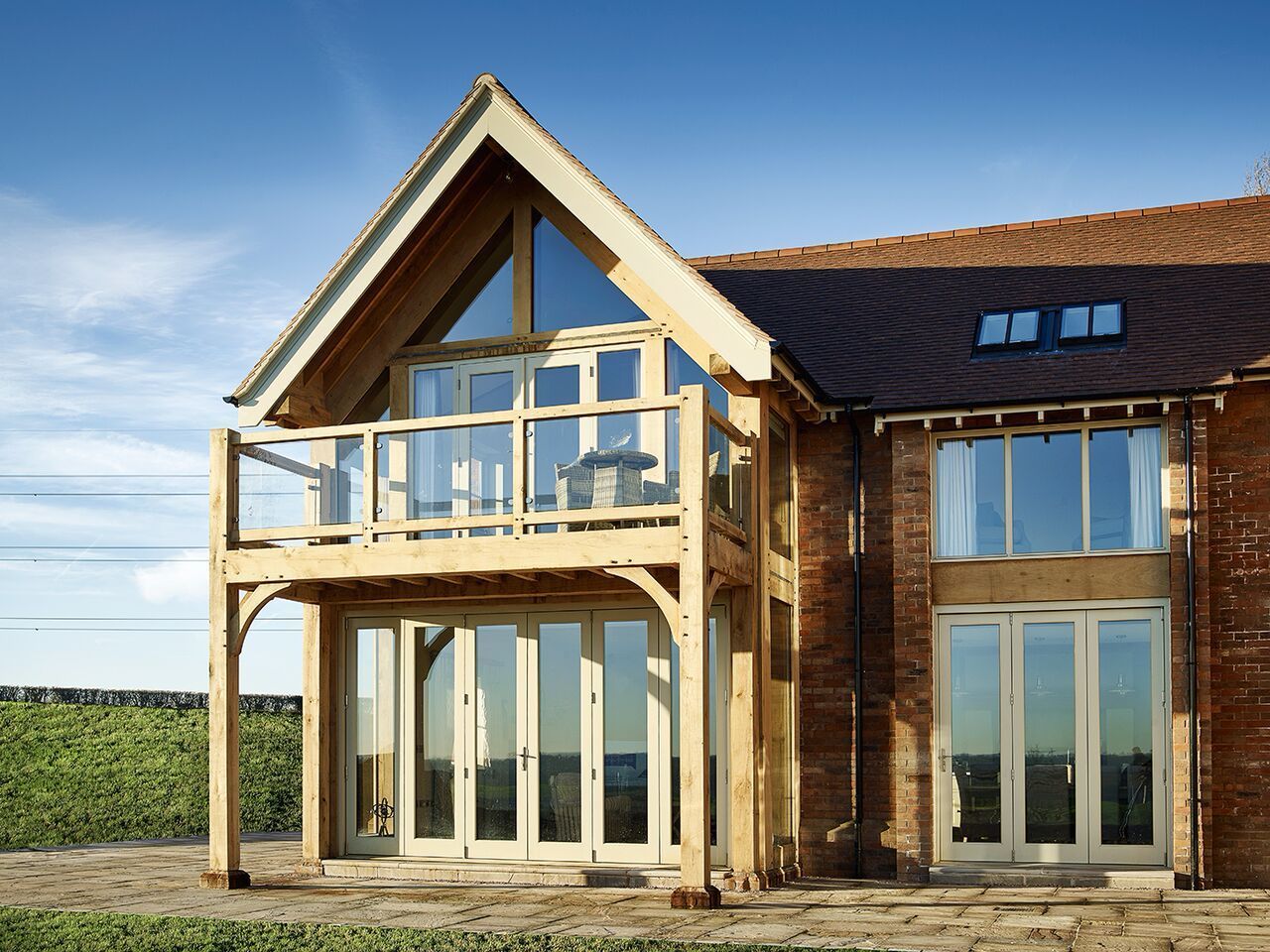 Two-story house with wood frame balcony, brick exterior, large windows, and a tiled roof against a blue sky.
