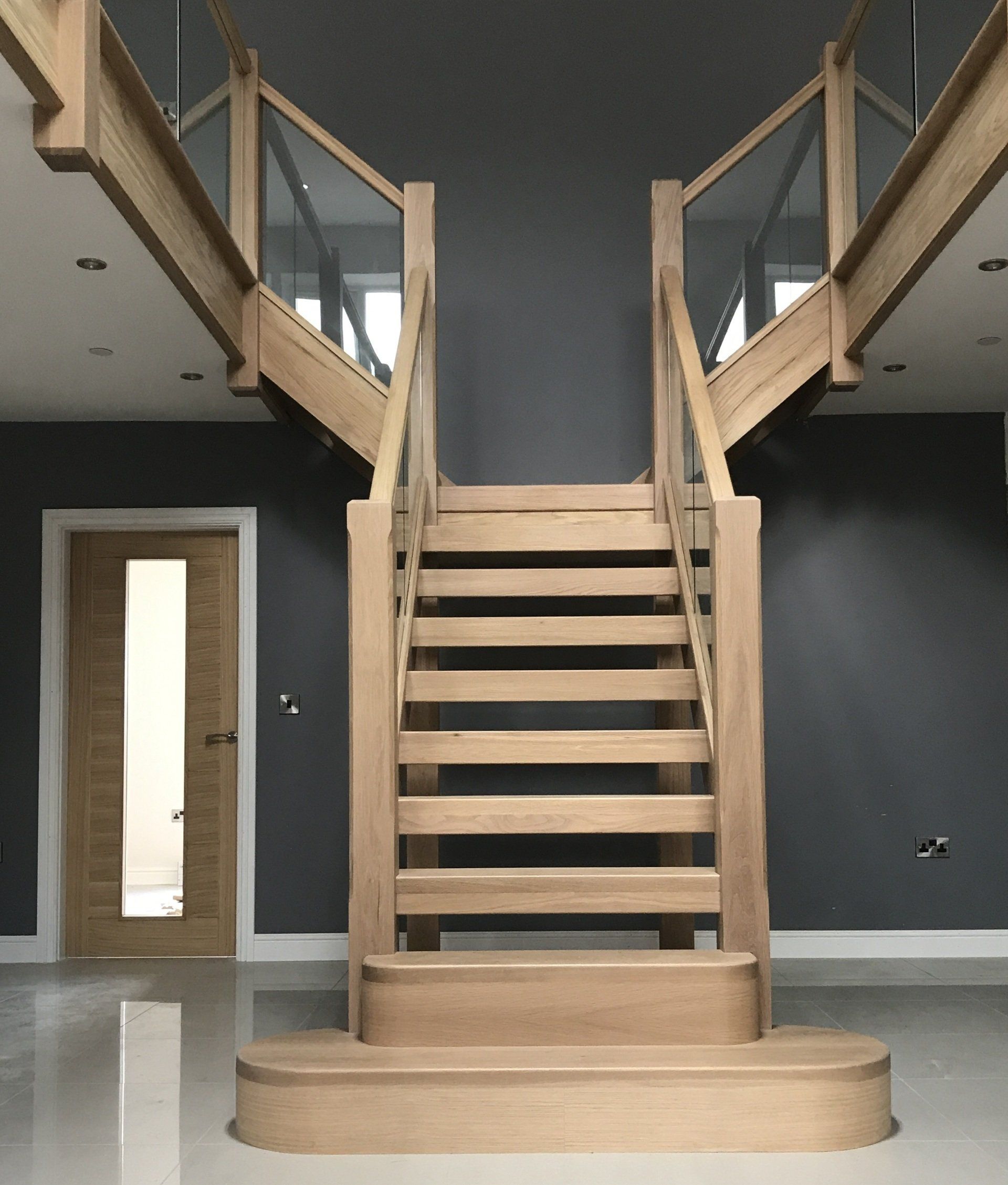 Wooden staircase with glass railings and a central divider, ascending to two landings, gray walls.