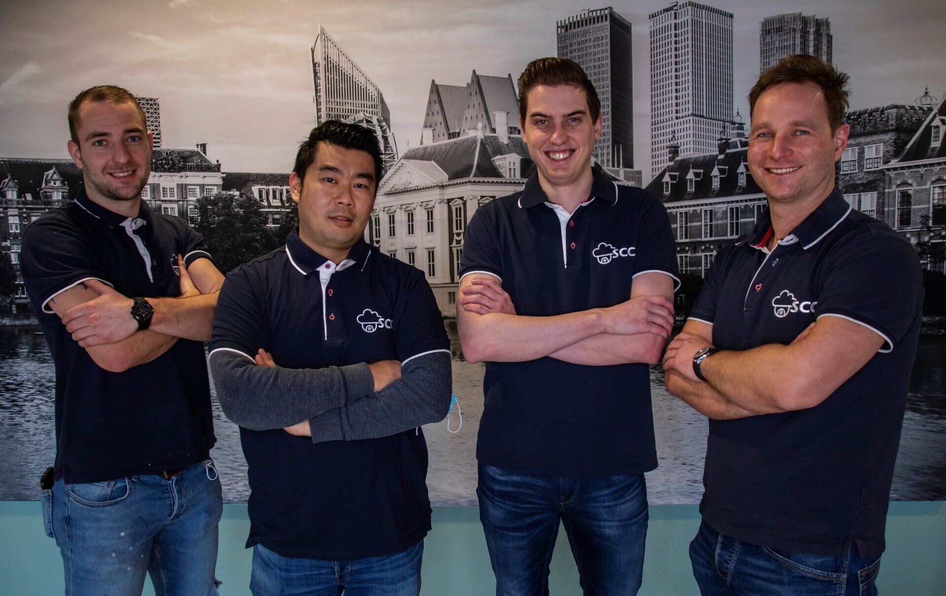 Vier mannen poseren voor een foto met hun armen gekruist voor de skyline van een stad.