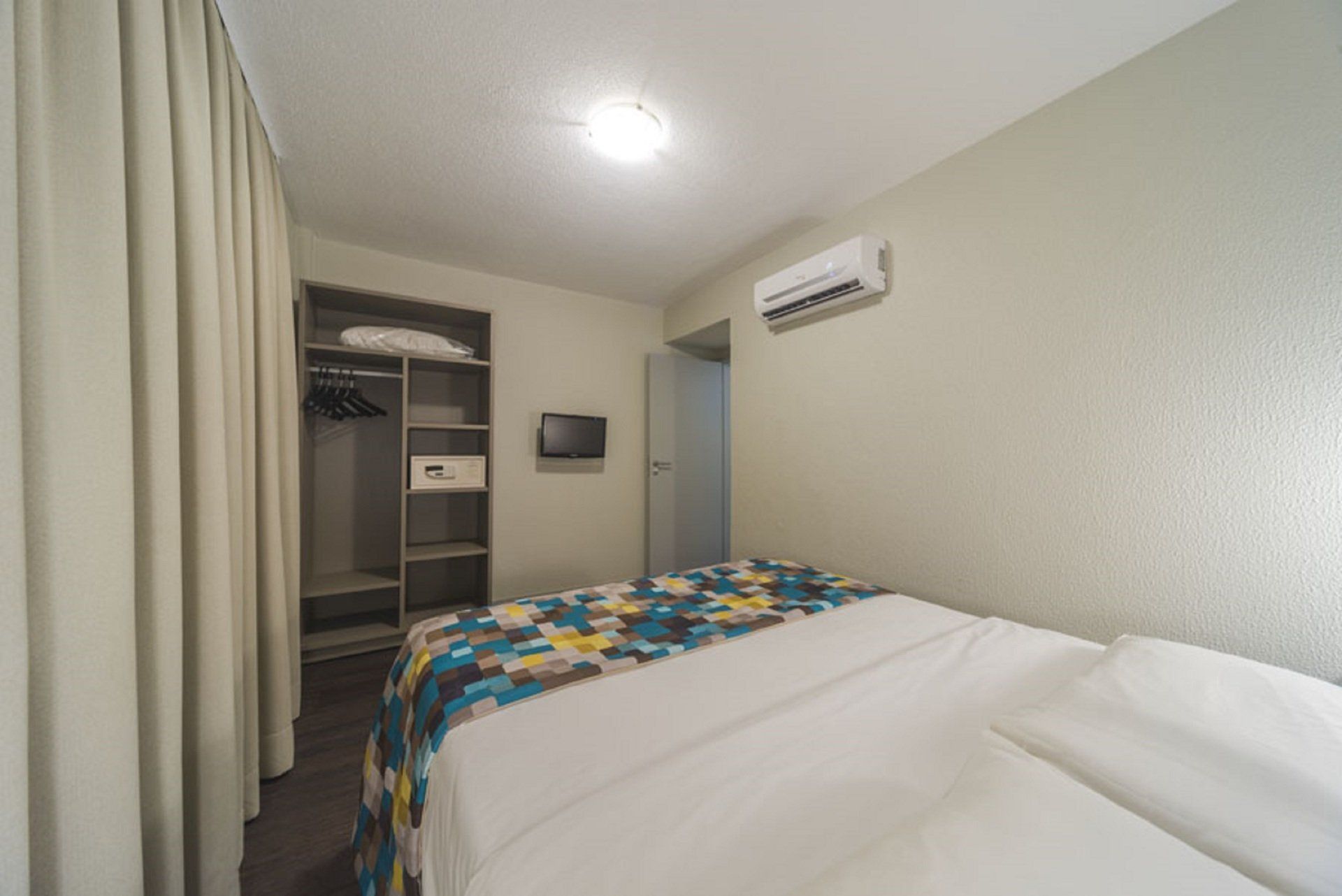 Quarto pequeno com cama, armário aberto, TV de parede e ar condicionado.