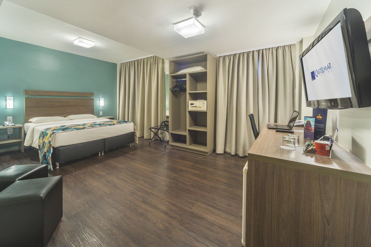 Quarto de hospital com cama, cadeira, TV, cortinas e piso de madeira.