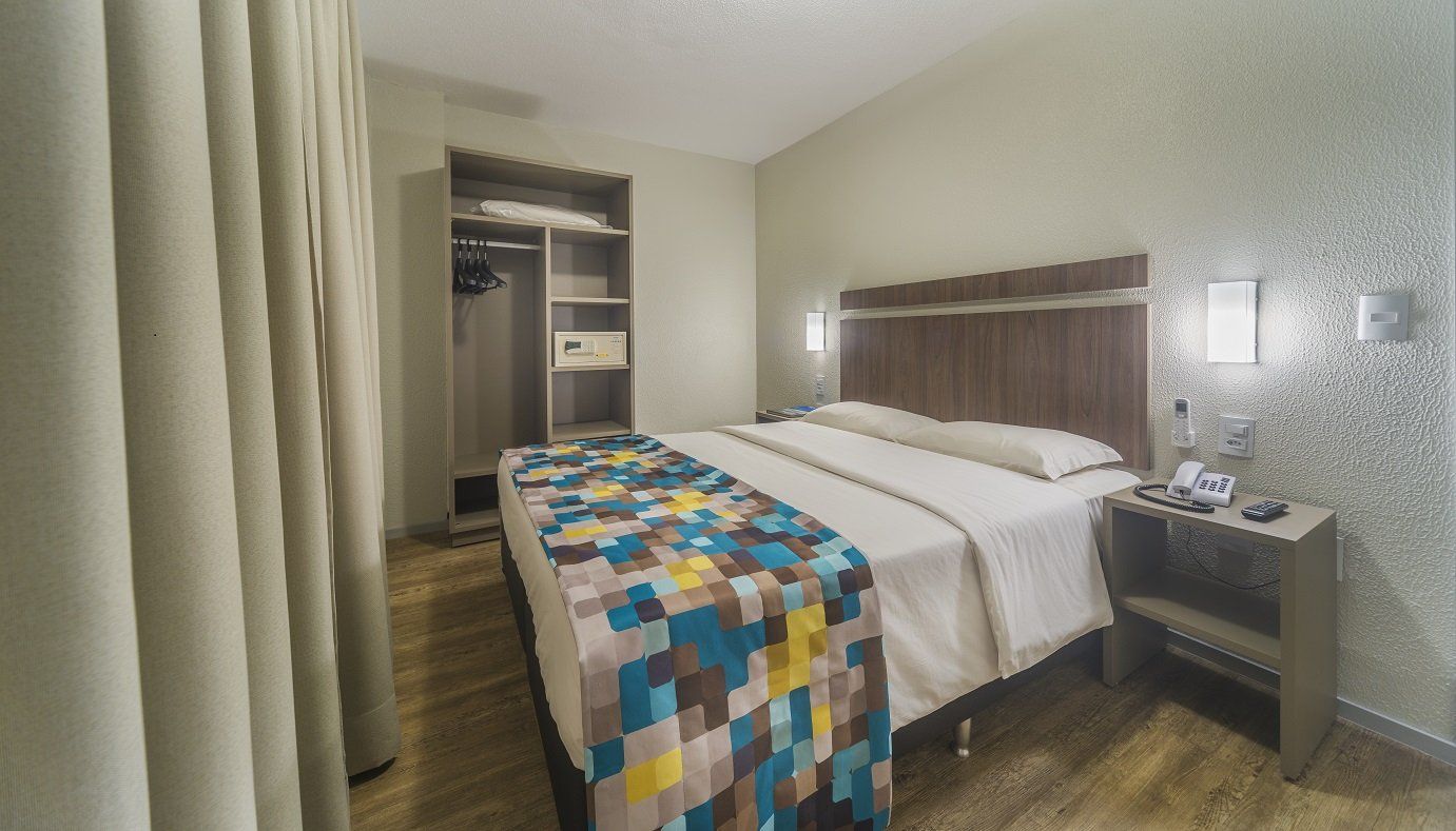 Quarto de hotel com cama, cobertor multicolorido, abajur de cabeceira e closet aberto.