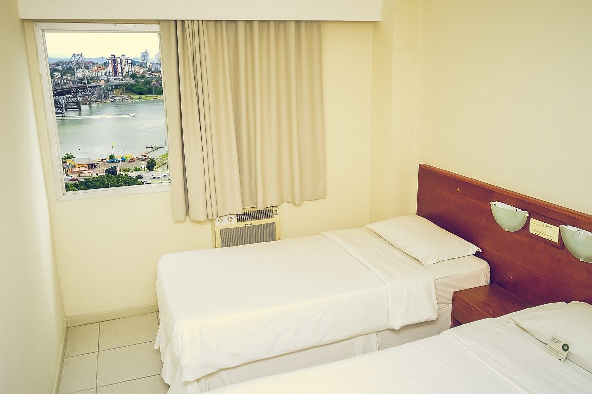 Quarto de hotel pequeno com duas camas brancas, cortinas bege e janela com vista para um rio.
