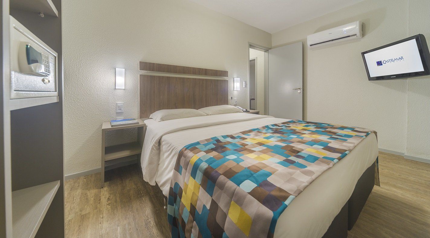Quarto de hotel moderno com cama grande, mesa de cabeceira, TV de parede e ar-condicionado.